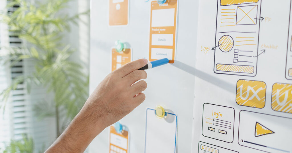 A hand holds a pen next to ui diagrams and sketches on a white board.